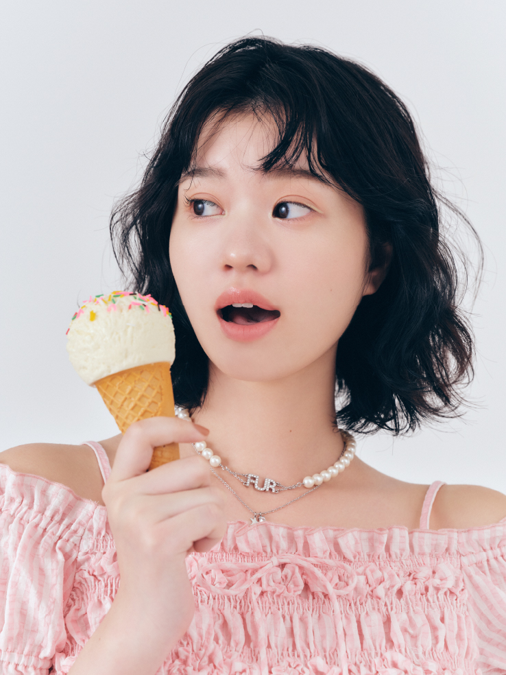 2WAYシャーリングブラウス・ロゴパールネックレスを着用した女性