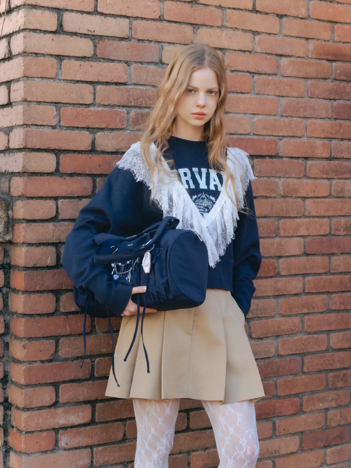 ベージュのフリルデザインテーラードジャケット・ベージュのベルト付きタックミニスカート・ネイビーのリメイクデザインフリルスウェット・ネイビーのリボンドラムバッグ・シルバーのロゴハートバッグチャーム・ブラウンのミニリボンカシメローファーを合わせたコーディネート