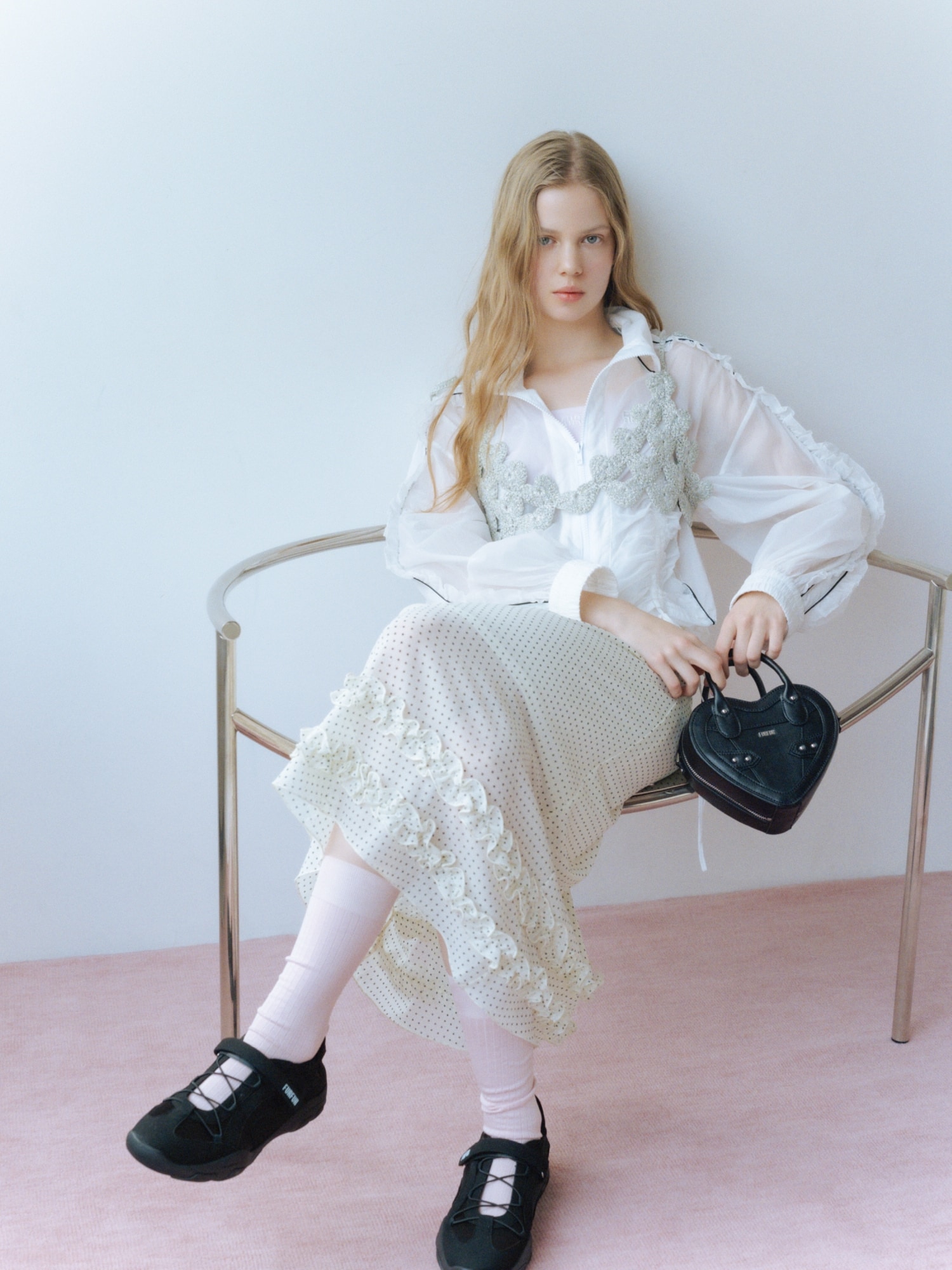 オフホワイトのシアーフリルブルゾン・白のミディナロースカート・シルバーのかぎ針ハートモチーフビスチェ・黒のハートショルダーバッグ・黒のメリージェーンデザインスニーカー・ピンクのロゴ刺繍バンドゥブラトップを合わせたコーディネート