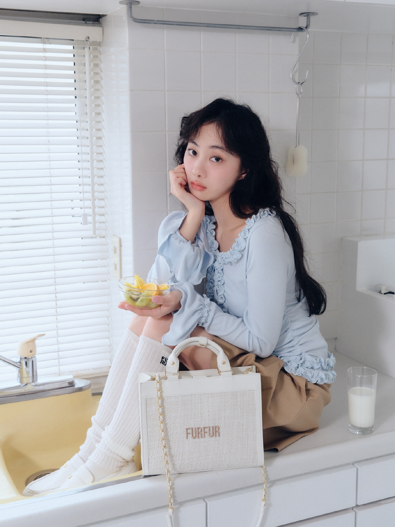 ライトブルーのフリルカーディガン・ベージュのベルト付きタックミニスカート・ベージュのフリルショートトレンチコート・アイボリーのツイードロゴトートバッグ・白のロゴシアーリブソックス・オフホワイトのミニリボンカシメローファーを合わせたコーディネート